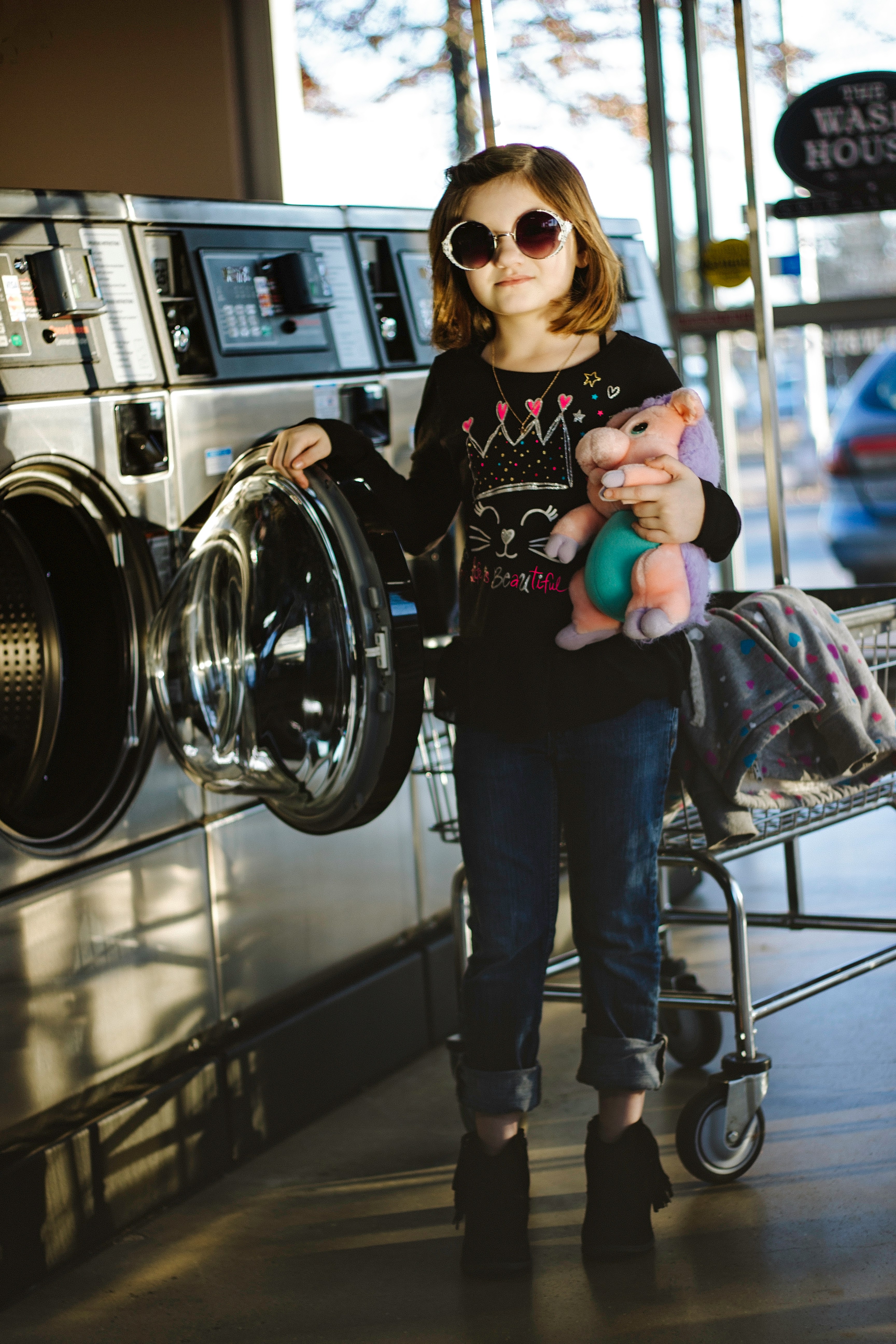 A girl aged about 6-7 is standing by the washing machine. She is about to put her teddy in it. She is wearing oversized sunglasses.