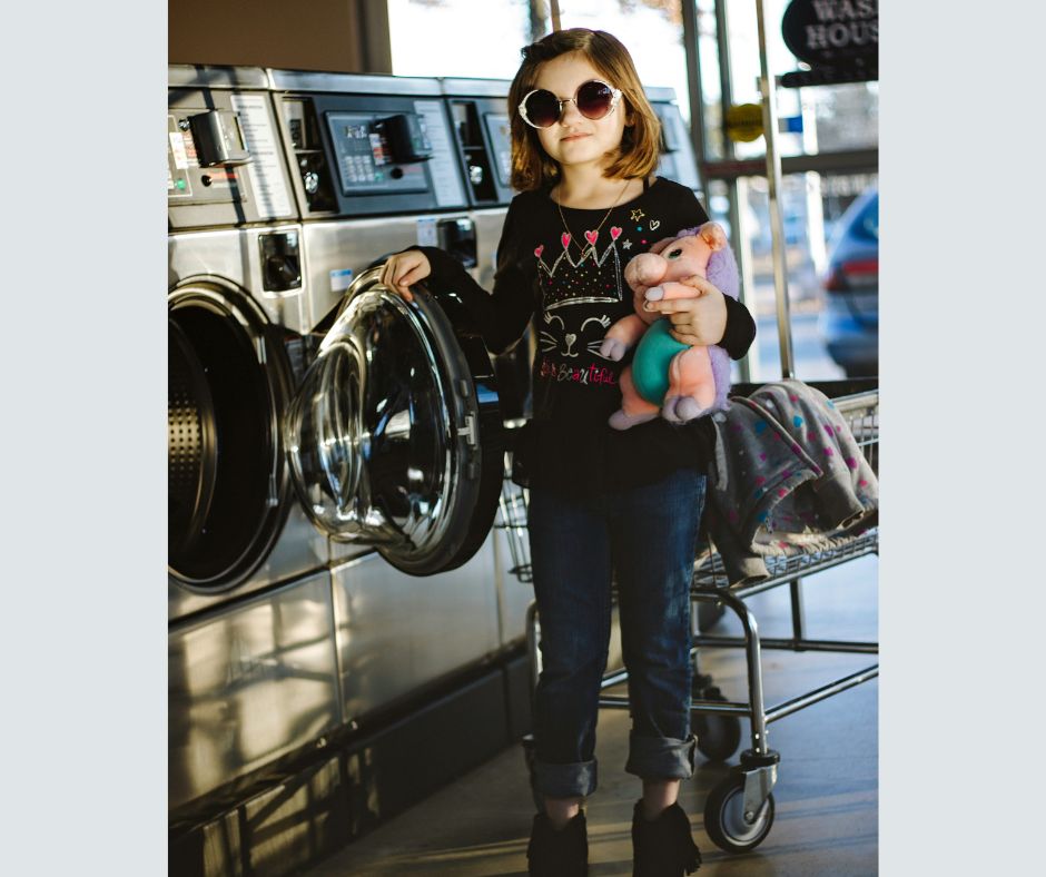 A 6-7-year-old girl is standing next to the washing machine. She is apparently about to put her teddy in it and wash it.