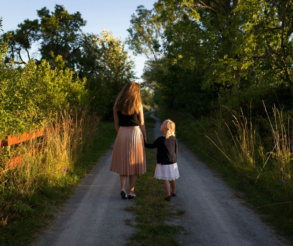 An image of a mother and daughter walking down the path holding hands. It is summer. They have their backs to us.