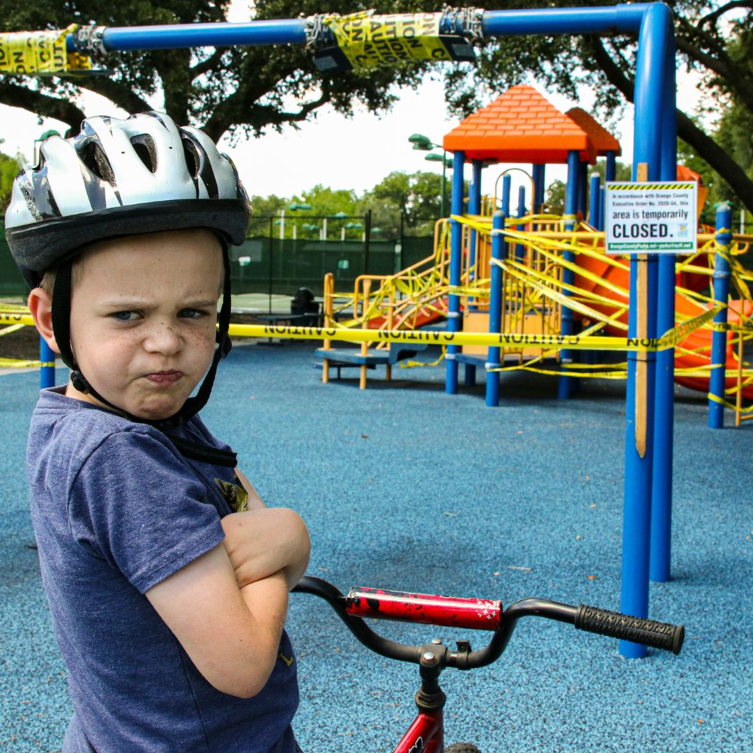 A boy of about 5 years of age looks displeased. His arms are crossed. He is wearing a bike helmet, standing in front of a closed playground.